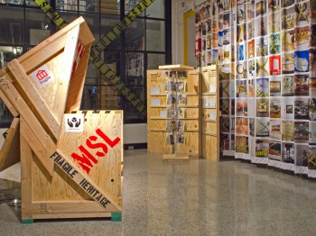 Gallery display with large wooden boxes and a hanging panel of fabric covered in square photos.