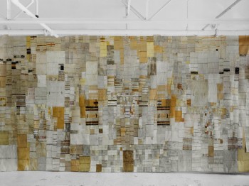 Large tapestry on a gallery wall made up of many small white, grey, tan, and orange rectangles.