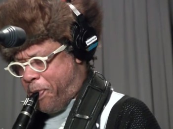 A man in a fur hat and headphones plays clarinet