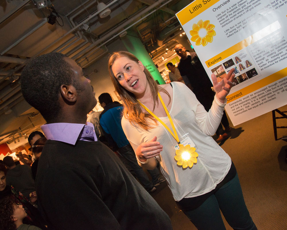 A woman wearing a sun-shaped lamp speaks in front of a poster.