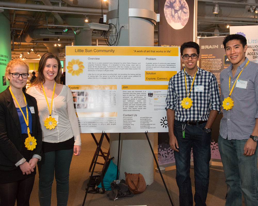 Four people wearing sun-shaped lights on lanyards pose next to a poster reading "Little sun Community"