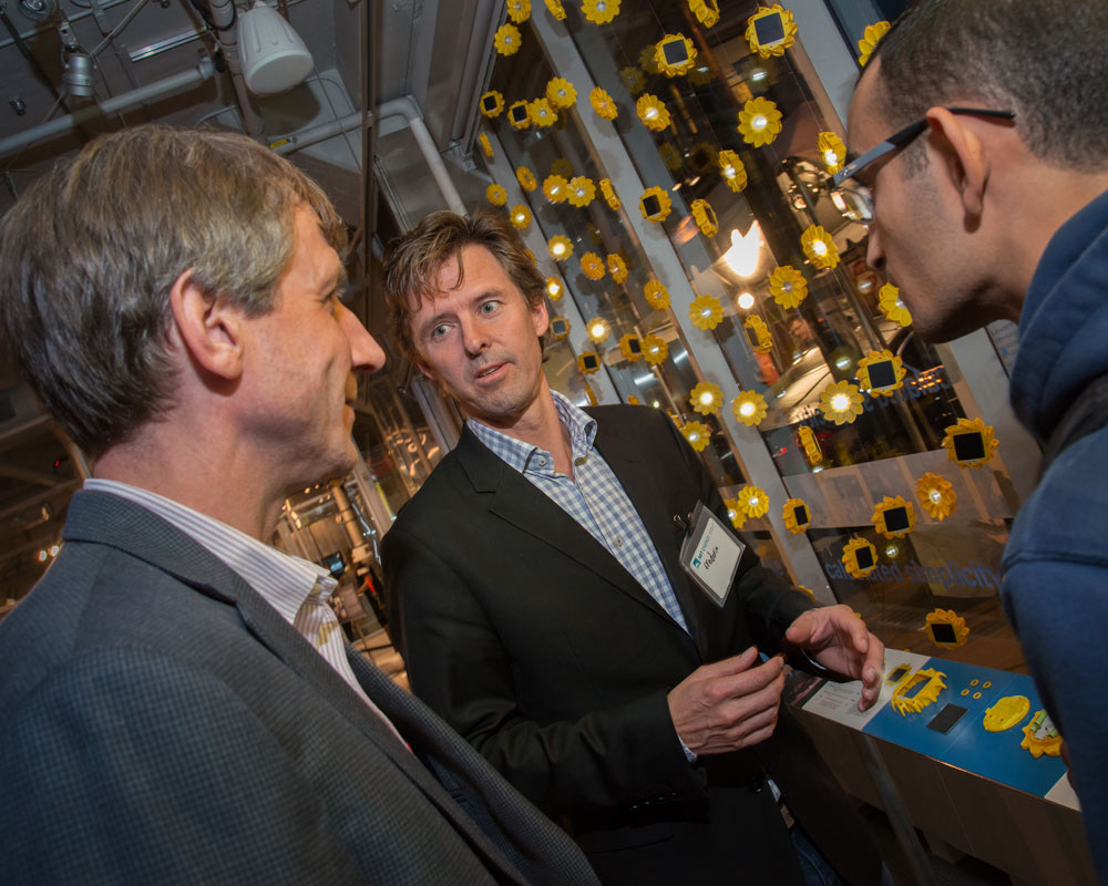 Three men in conversation in front of a display of many small sun-shaped lamps.