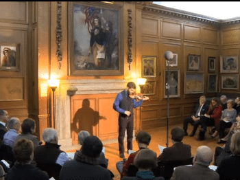 A man performs violin for an audience in a historic gallery