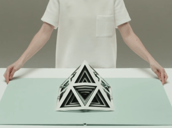 A person stands behind a pyramid-shaped sculpture made of triangles cut out from paper.