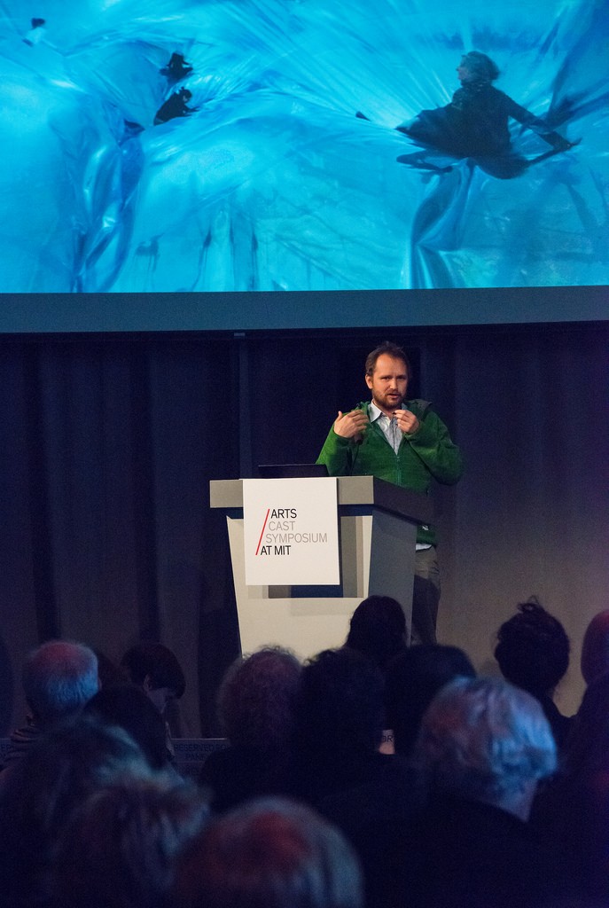 A man speaks at a podium. An image of people playing on large plastic sheets is projected behind him.