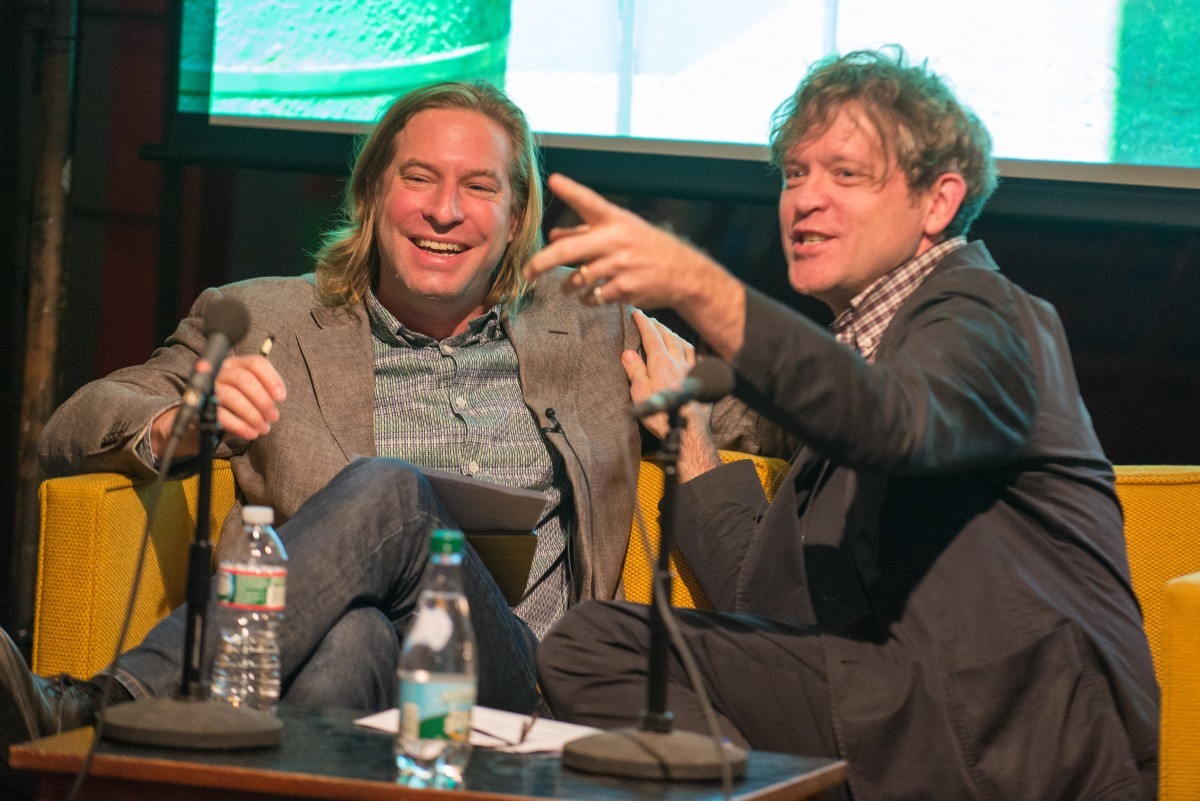 A man laughs and a man points while in discussion on stage.