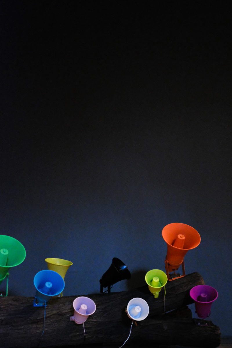 Installation view of eleven multicolored speakers attached to a tree laying on a hardwood floor.