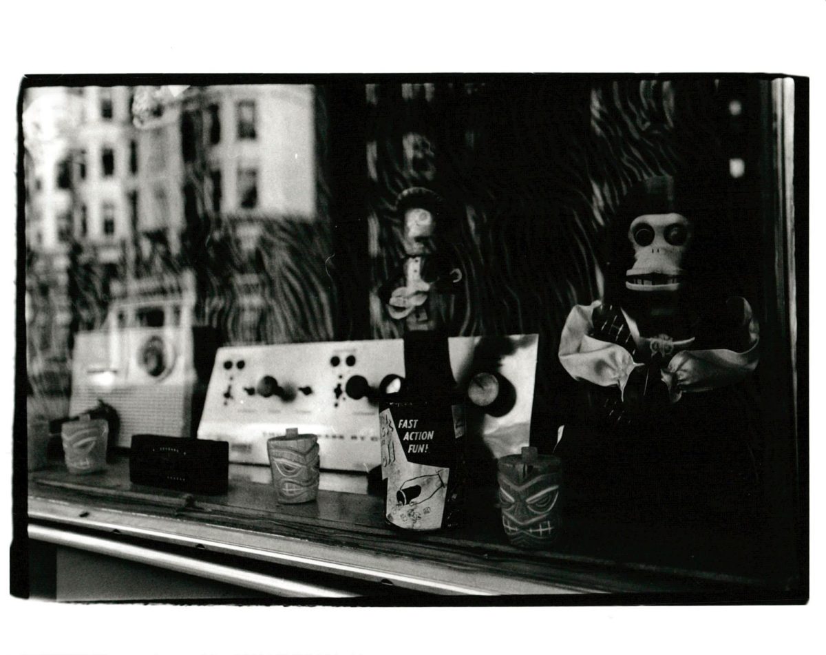 Black and white gelatin silver print looking into a store window display of several vintage items with a white three story building reflected in the window.