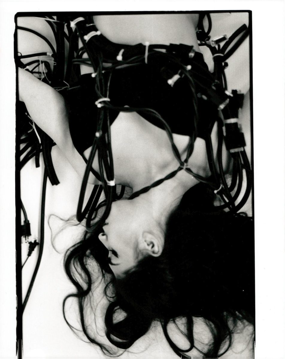 A figure lays on the ground with their long dark hair spread on the floor among a series of electric cables.