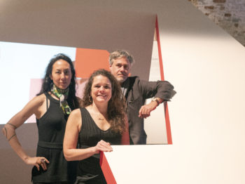 Three people pose next to a geometric architectural installation.