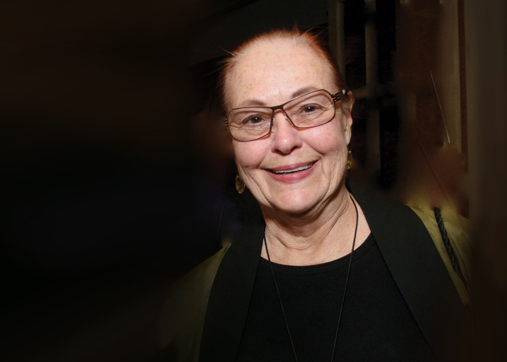 Portrait of Brit d’Arbeloff, facing camera and smiling in front of a dark background.