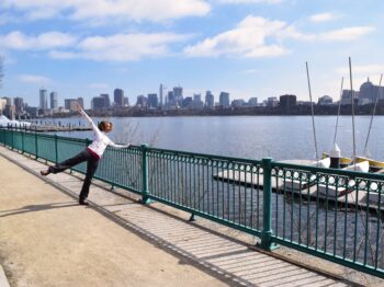 Adesola Akinleye stands in front of the Charles River and Boston Skyline with an arm and a leg raised.