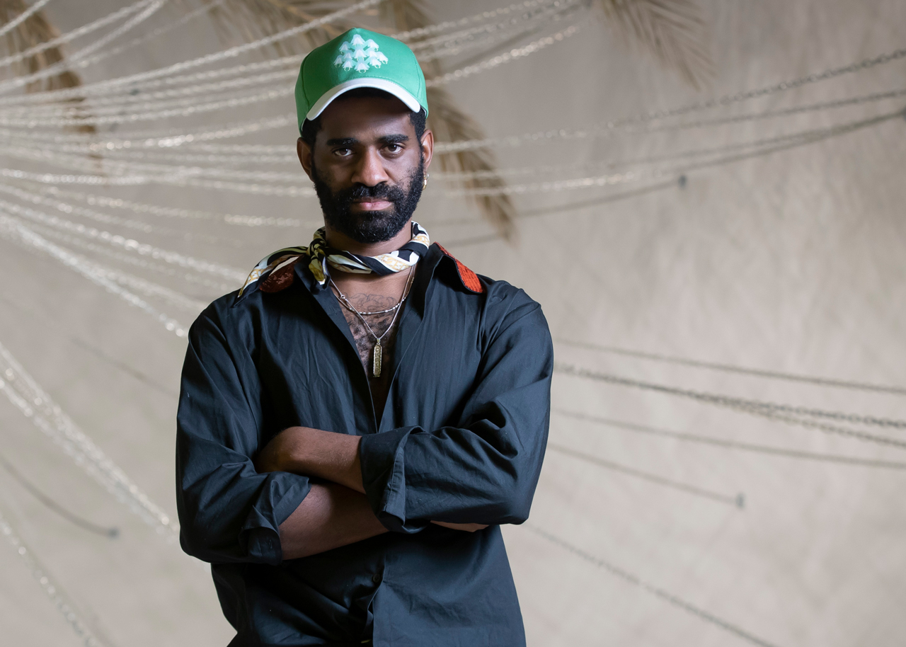 Christopher Joshua Benton looks directly into the camera with arms crossed, while standing in front of an artwork with spokes of lines radiating from the left side of the frame.