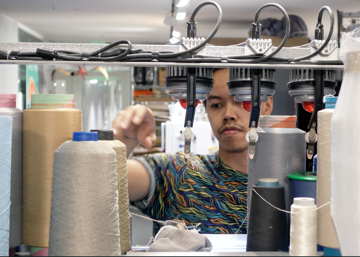 Irmandy Wicaksono surrounded by large spools of thread as he works with a digital knitting machine.