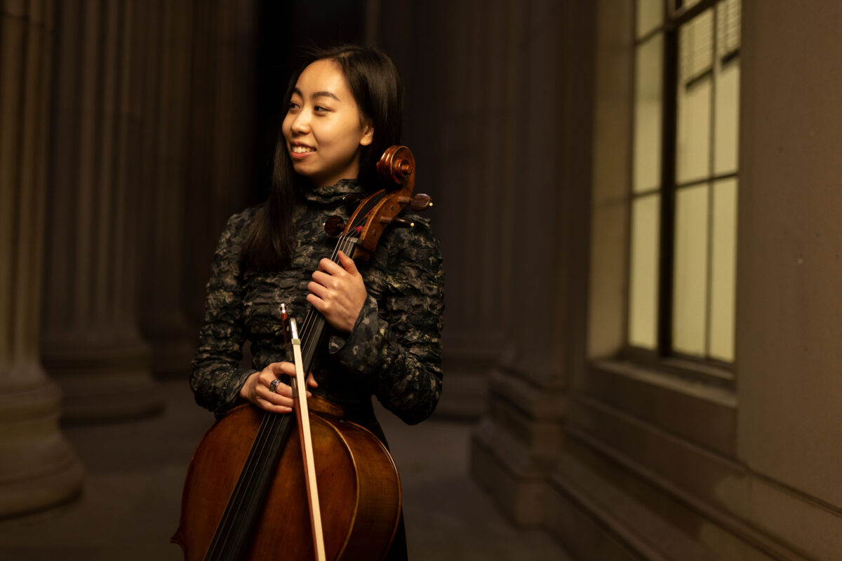 Woman in a hall holding a cello and looking to her right while smiling. 