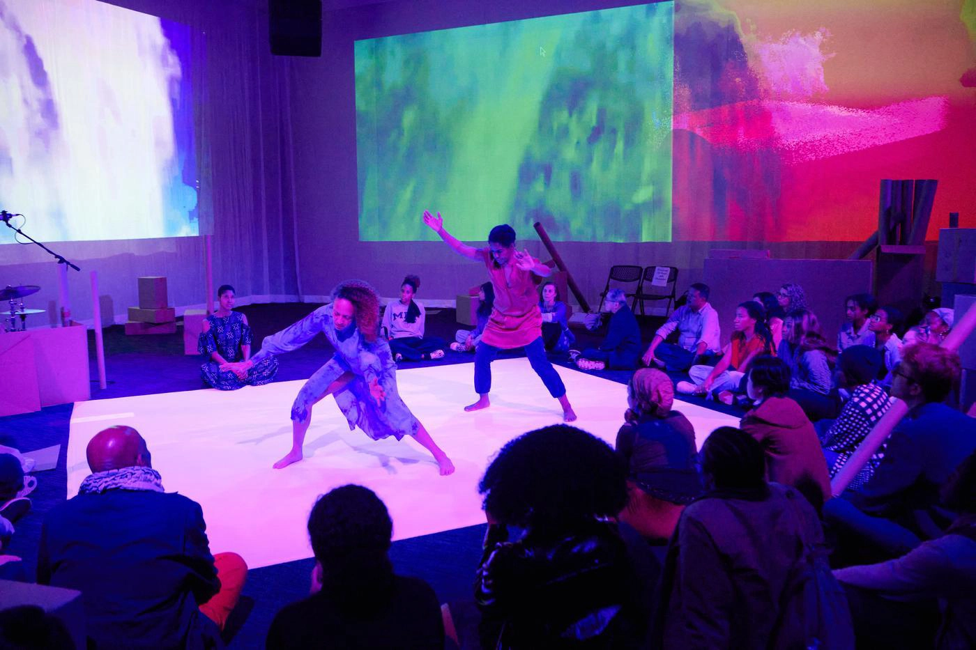 Two people performing at a stage in the center of a. crowed room colorfully lighted by projections on each wall.